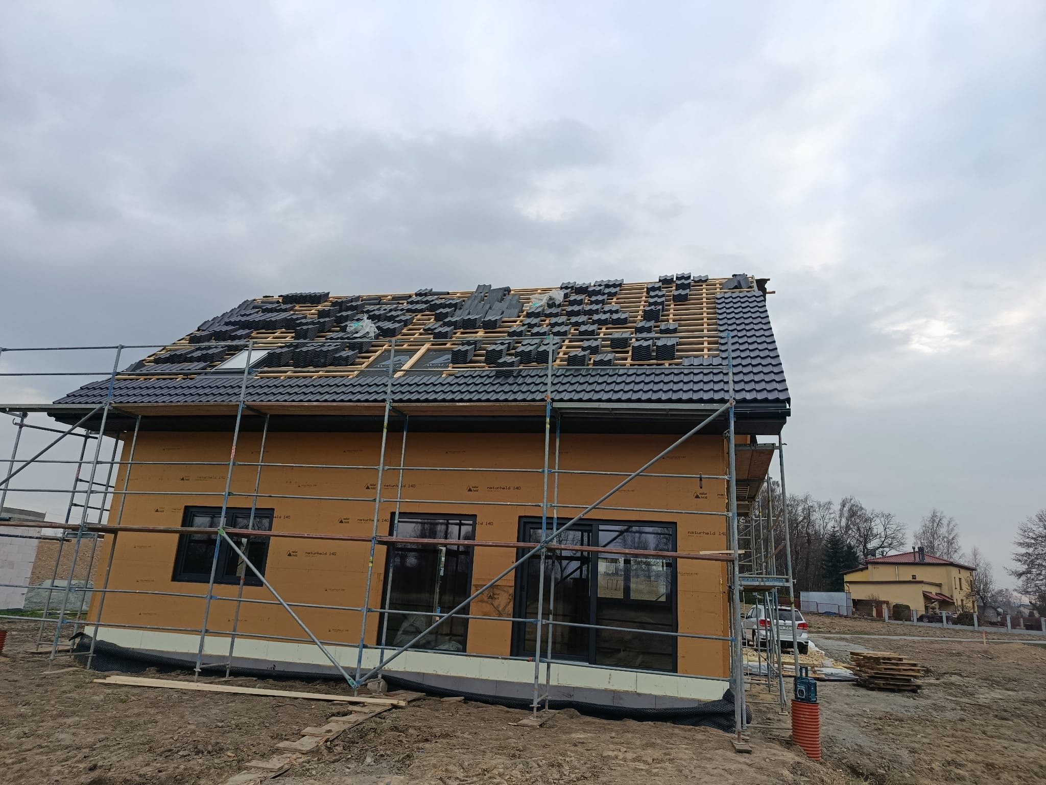 Budowa domu w trakcie: widoczne ściany z płyt, rusztowania i częściowo pokryty dach z czarną dachówką. Niebo pochmurne, w tle inne budynki.