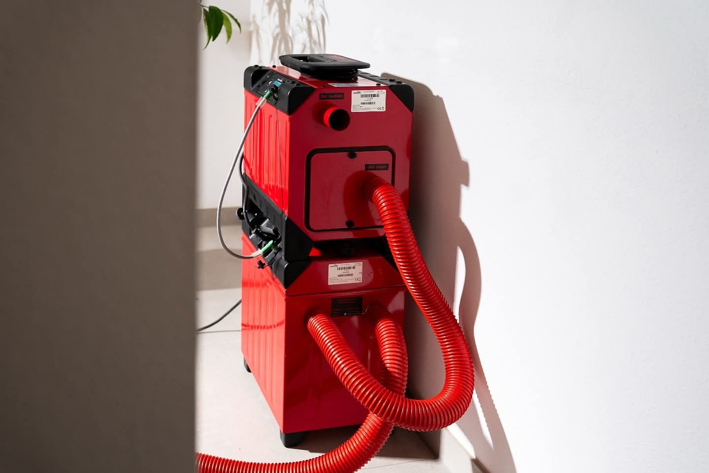 Czerwony, dwuczęściowy kondensacyjny osuszacz powietrza z wężami, stojący przy białej ścianie, oświetlony naturalnym światłem. Widoczne etykiety i złącza.