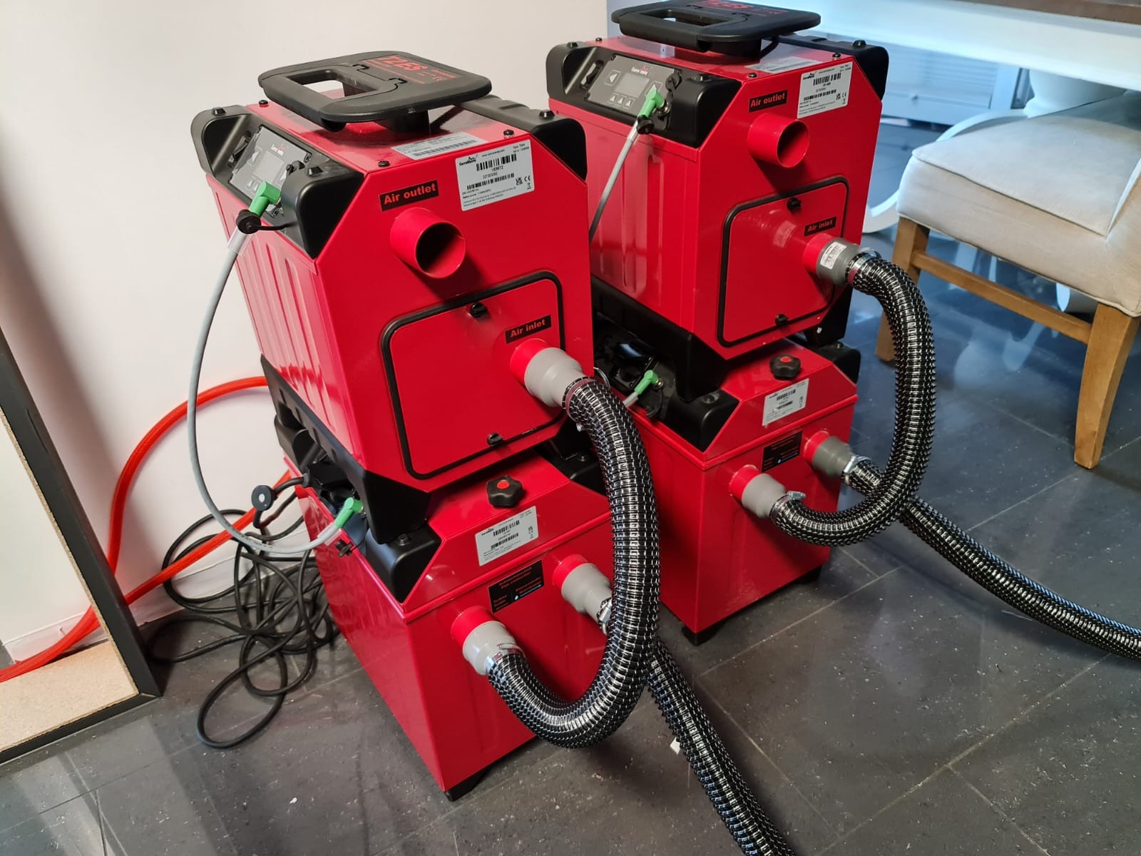 Dwa czerwone, profesjonalne urządzenia do osuszania z podłączonymi wężami, ustawione na ciemnej podłodze w pomieszczeniu. Widoczne detale: wloty i wyloty powietrza.