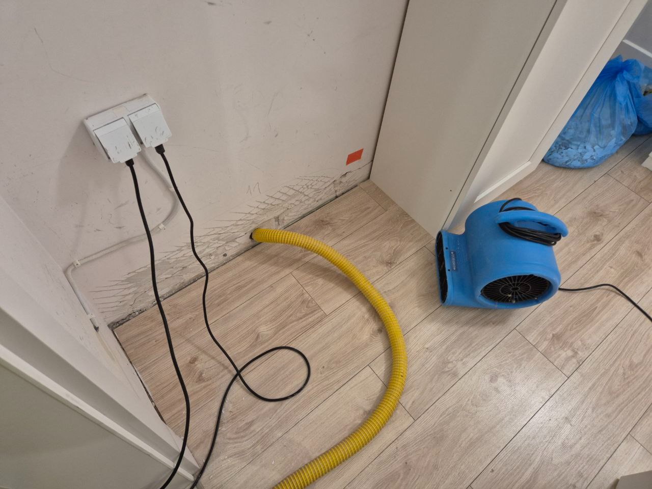 Niebieski osuszacz podłączony do ściany z widocznymi śladami wilgoci i gniazdkiem elektrycznym. Żółty wąż doprowadzony do ściany. Widoczne ślady uszkodzeń po zalaniu.