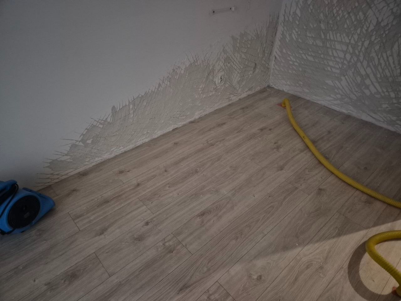 Pokój po zalaniu z widocznymi śladami wilgoci na ścianach i podłodze. Żółty wąż i niebieskie urządzenie do usuwania wilgoci w rogu pomieszczenia.