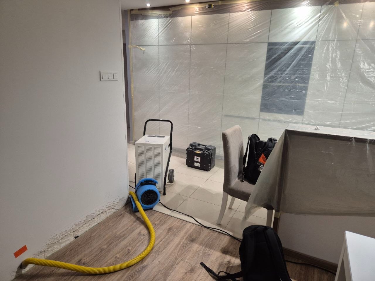 Pokój zabezpieczony folią malarską z urządzeniem do usuwania wilgoci i żółtym wężem. Widoczne uszkodzenia ściany przy podłodze.