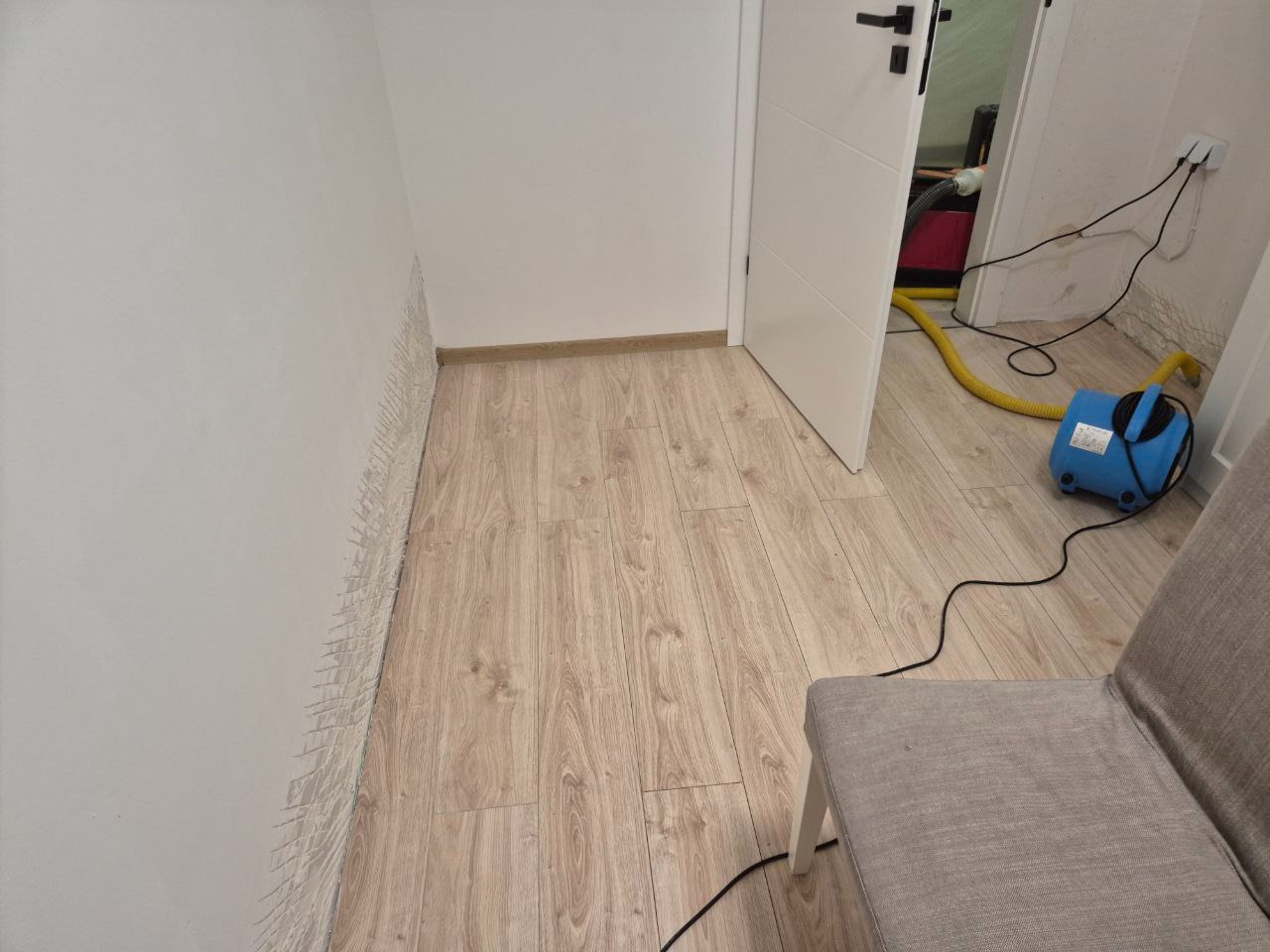 Narożnik pokoju z widocznymi śladami wilgoci na ścianie, obok stoi niebieski wentylator z żółtym wężem, skierowany w stronę otwartych drzwi. Podłoga wyłożona panelami.