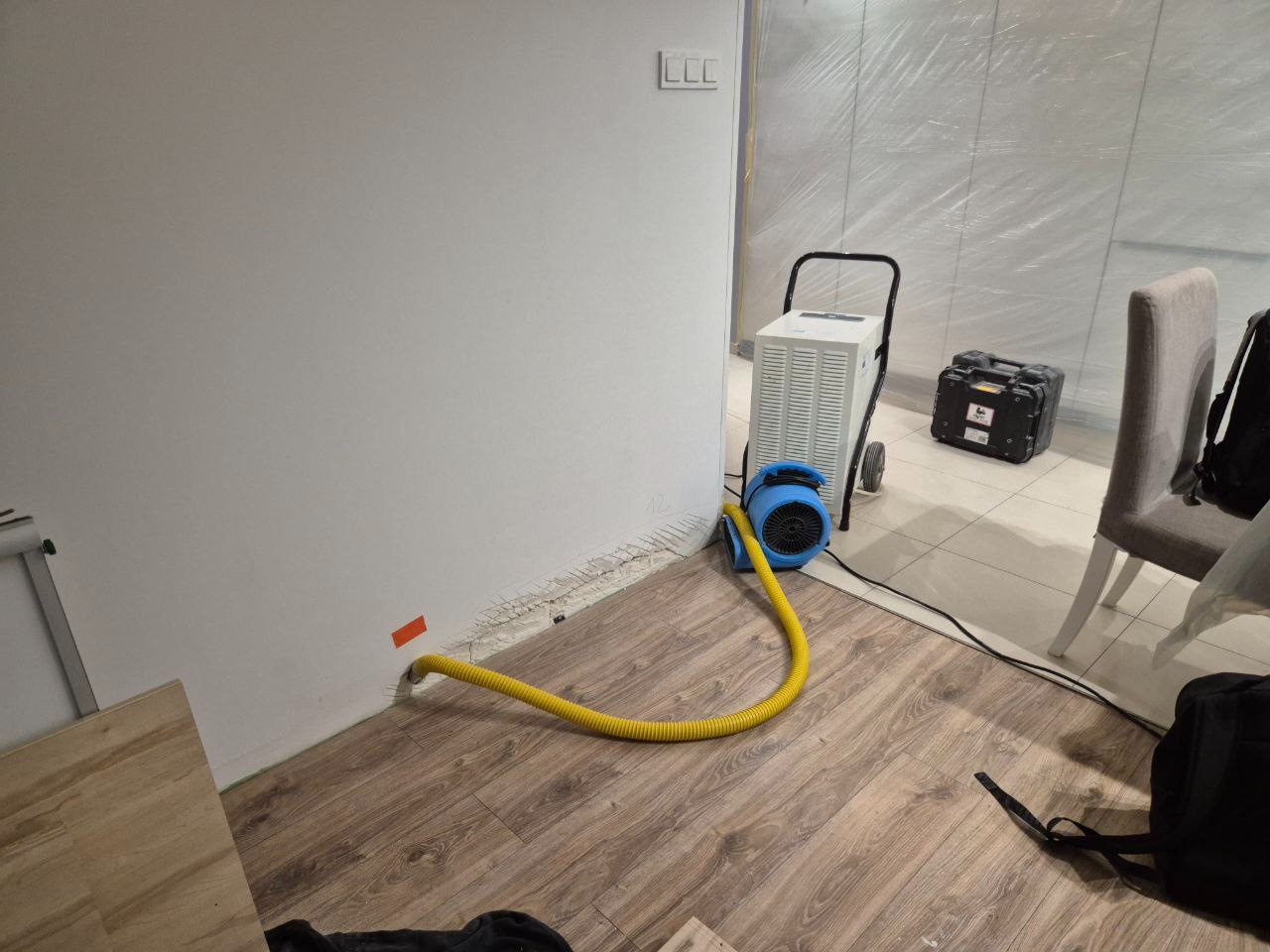 Biała ściana z żółtym wężem podłączonym do urządzenia na kółkach. Widoczne ślady wilgoci przy podłodze i folia ochronna na ścianach w tle. Sprzęt do usuwania wilgoci.