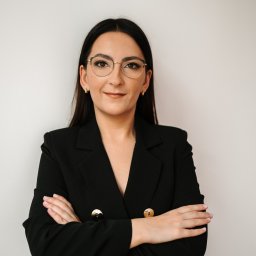 Magdalena Kubat Biuro Rachunkowe - Portret kobiety w czarnej marynarce z ozdobnymi guzikami, skrzyżowane ręce, okulary w złotych oprawkach, na jasnym tle. Profesjonalny wizerunek.