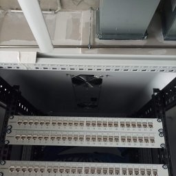Łukasz Bonikowski ELEKTRO SERWIS - Panel krosowniczy w szafie rack, widoczne porty RJ45 z numeracją, wentylator chłodzący, sufit z instalacjami. Ujęcie z dołu, oświetlenie techniczne.
