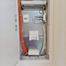 Łukasz Bonikowski ELEKTRO SERWIS - Otwarta skrzynka rozdzielcza z uporządkowanymi kablami (pomarańczowe i białe) oraz gniazdami. Widoczne elementy instalacji elektrycznej i teletechnicznej wewnątrz obudowy.