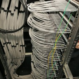 Łukasz Bonikowski ELEKTRO SERWIS - Uporządkowane kable w serwerowni, ułożone w wiązki i przymocowane do metalowej ramy. Widoczne oznaczenia na kablach i fragment dłoni w prawym górnym rogu.