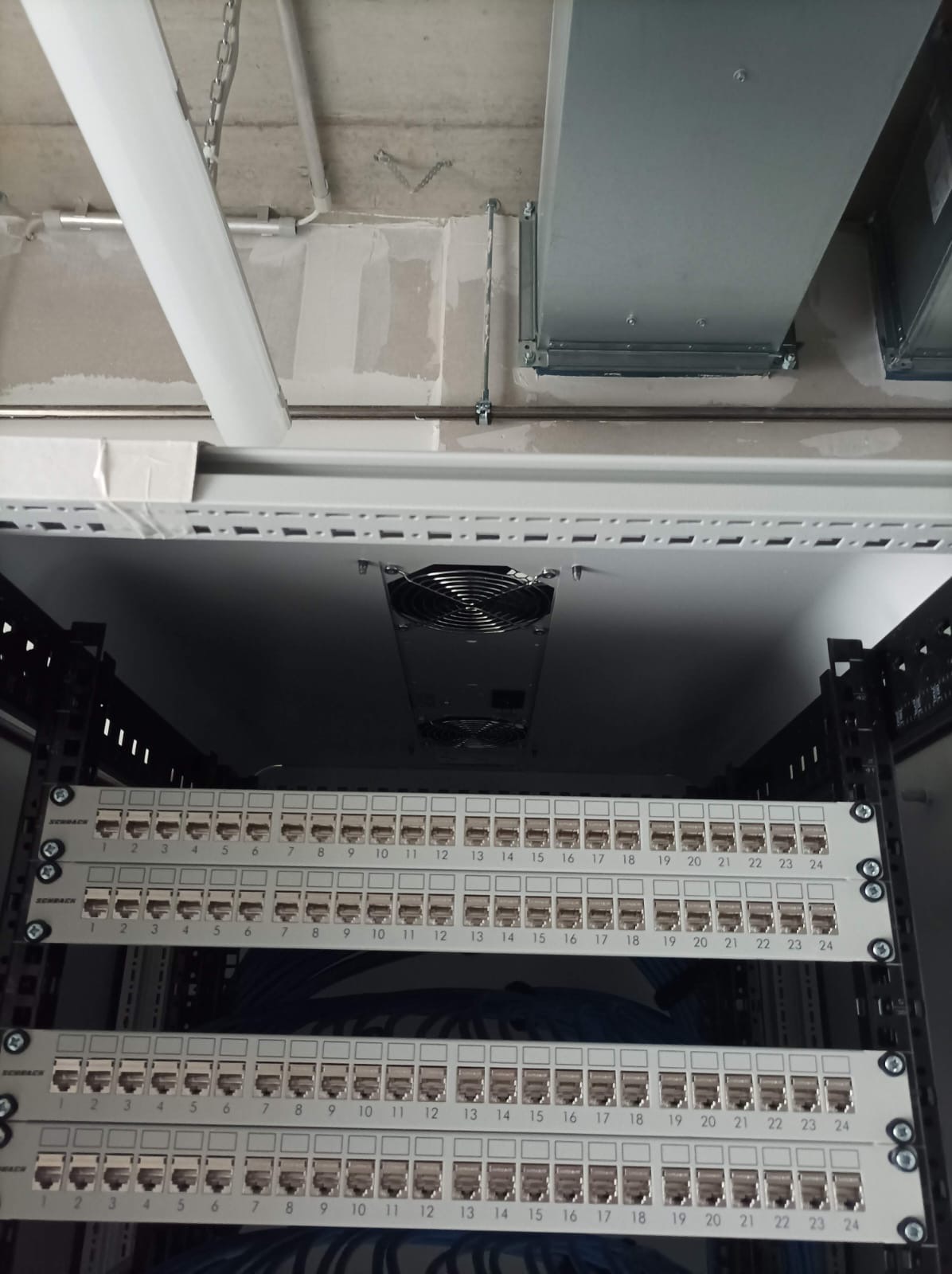 Panel krosowniczy w szafie rack, widoczne porty RJ45 z numeracją, wentylator chłodzący, sufit z instalacjami. Ujęcie z dołu, oświetlenie techniczne.