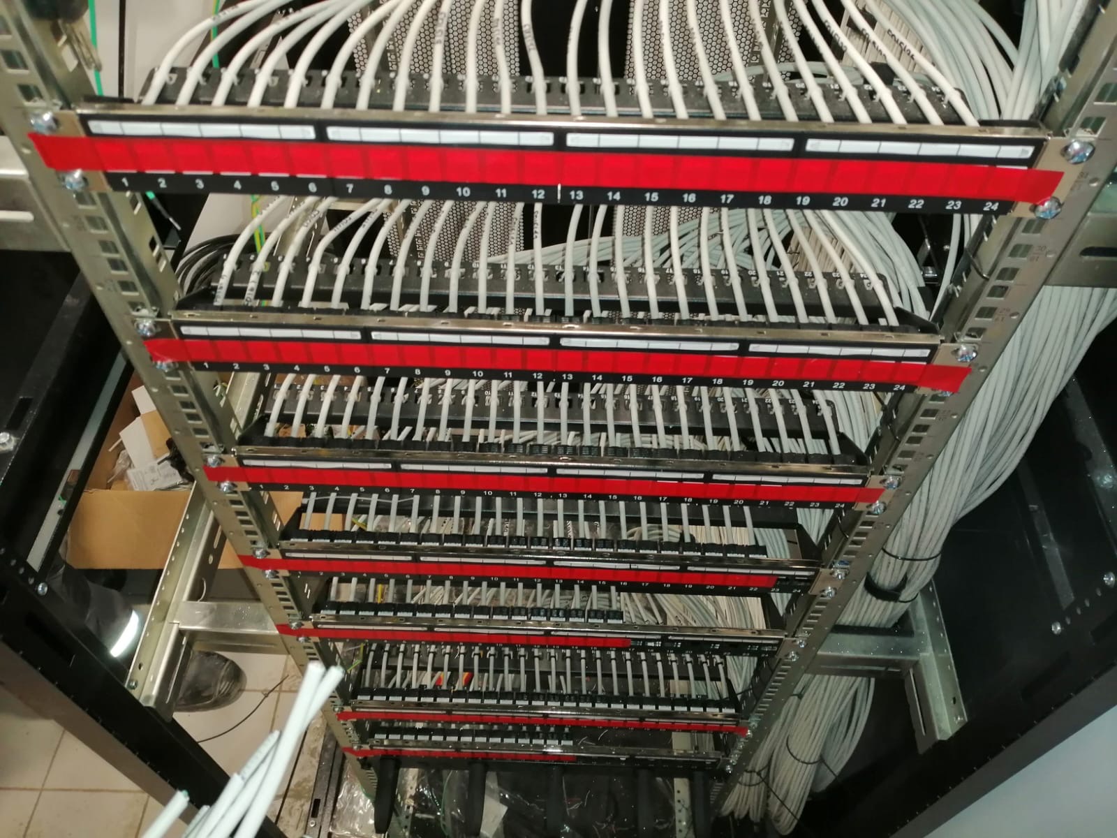 Panel krosowniczy z podłączonymi kablami sieciowymi, oznaczony numerami od 1 do 24, zamontowany w metalowej szafie rack. Widok z góry, perspektywa z dołu.