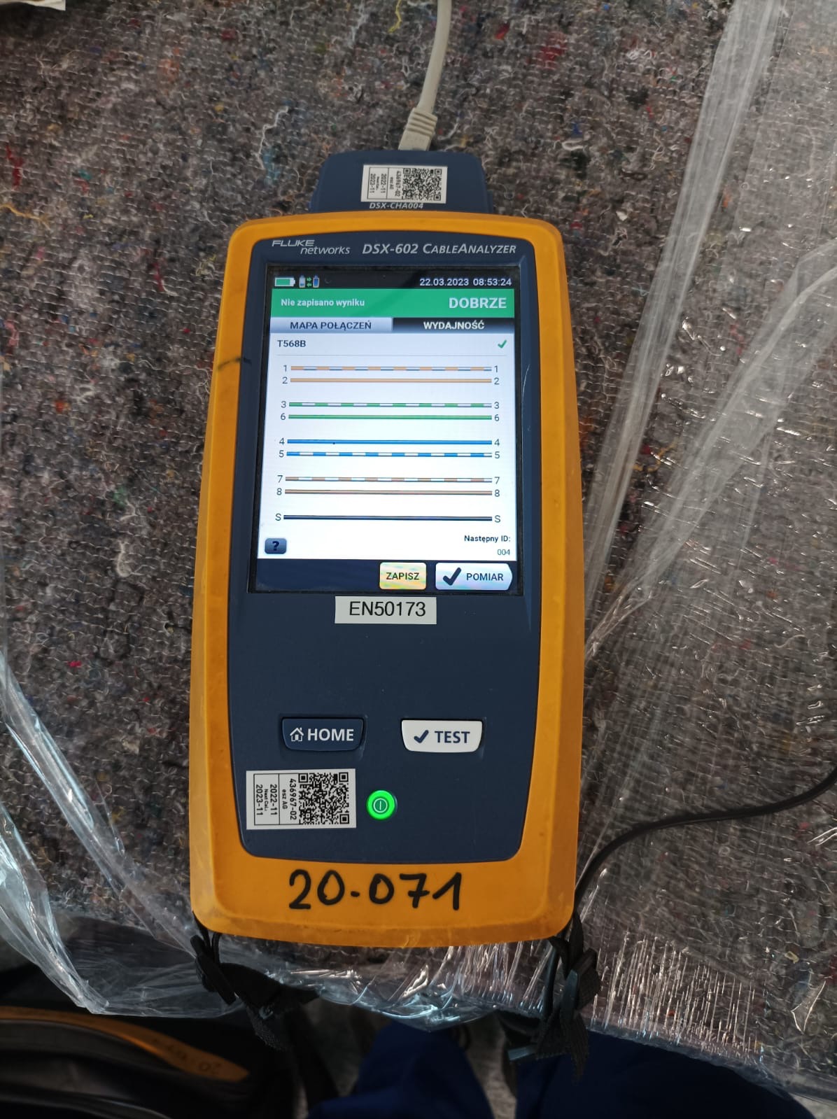 Tester okablowania Fluke DSX-602 z pozytywnym wynikiem testu T568B, leżący na szarym materiale. Widoczny kabel podłączony do urządzenia i numer identyfikacyjny 20-071.