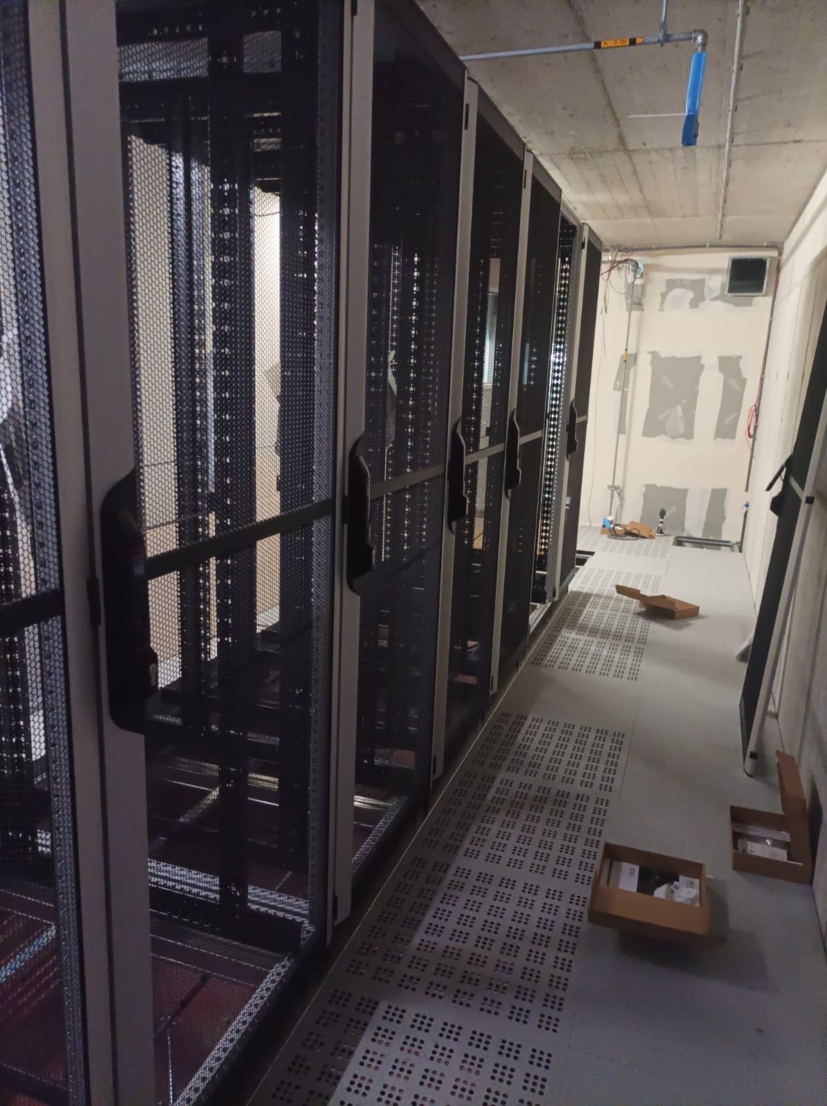 Rząd metalowych szaf serwerowych z perforowanymi drzwiami w surowym pomieszczeniu technicznym z widocznymi instalacjami pod sufitem i podłogą.