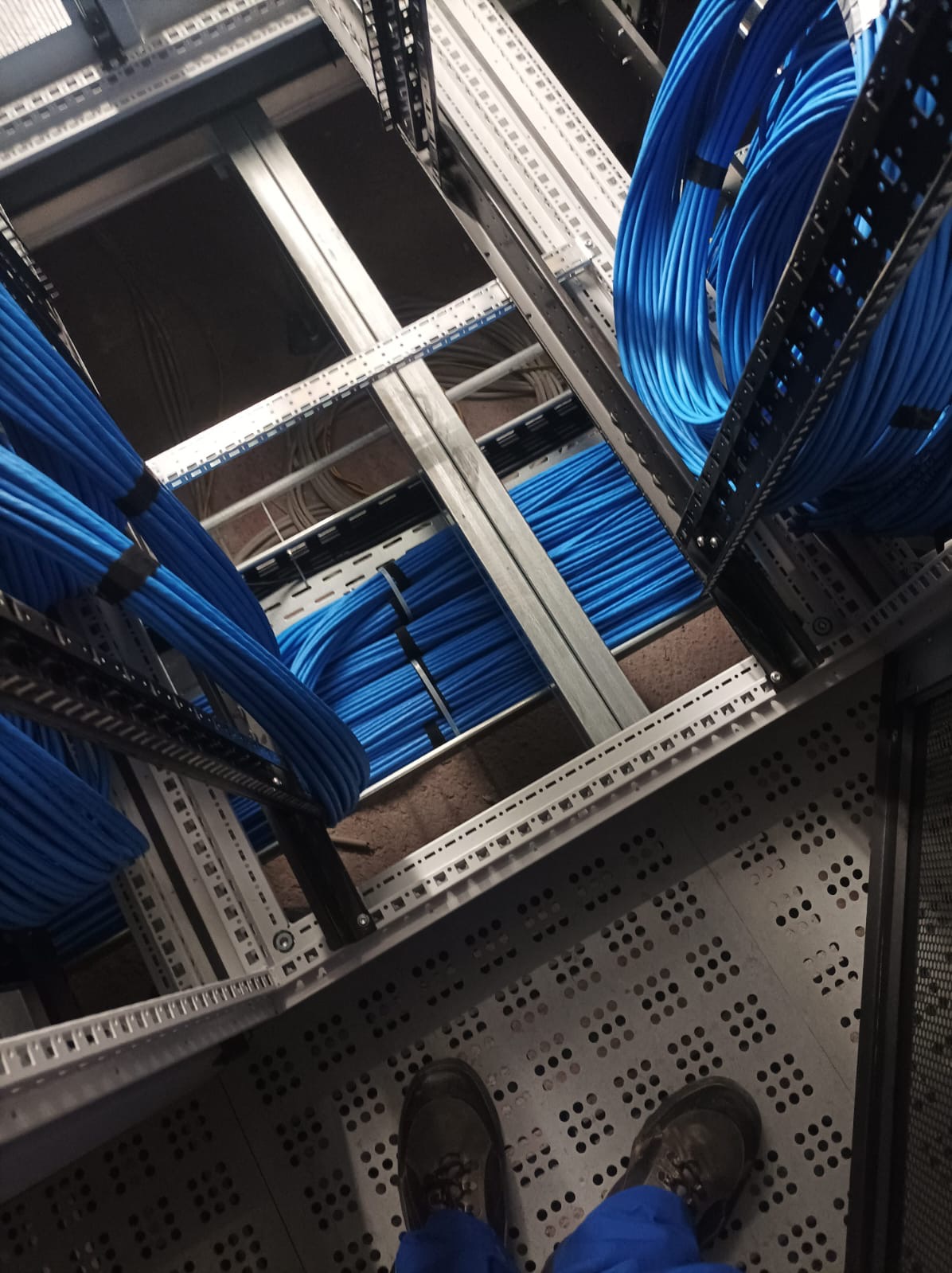 Ułożone niebieskie kable w metalowej konstrukcji, widok z góry na stopy osoby w kombinezonie roboczym. Kable równo ułożone, zabezpieczone opaskami.