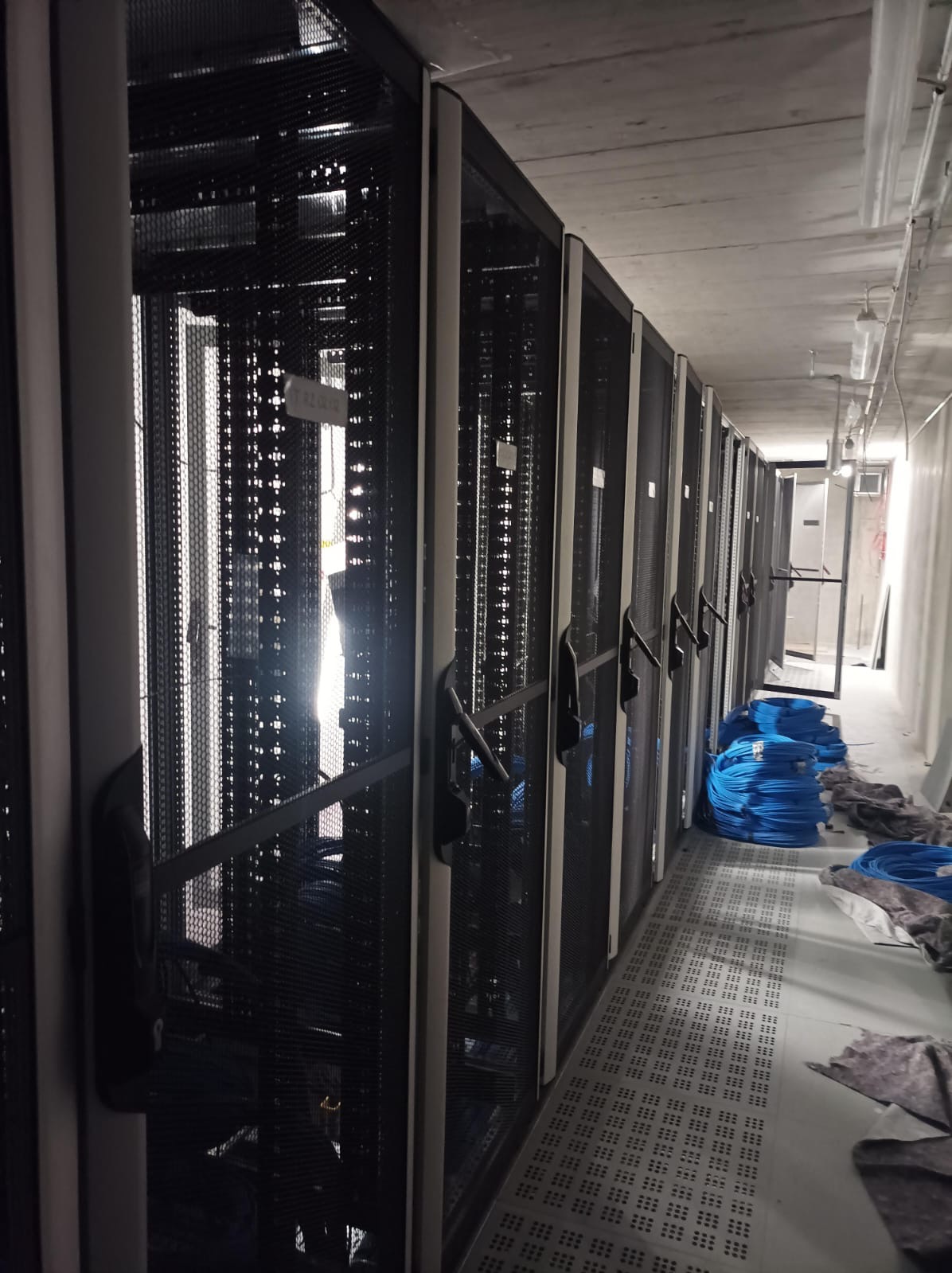 Rząd szaf serwerowych w serwerowni, widoczne niebieskie kable i kratka podłogowa. Oświetlenie sufitowe. Metalowe szafy z perforowanymi drzwiami.