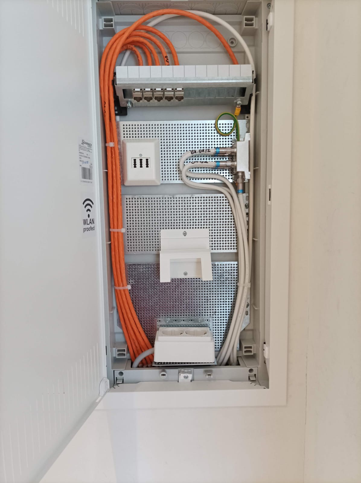 Otwarta skrzynka rozdzielcza z uporządkowanymi kablami (pomarańczowe i białe) oraz gniazdami. Widoczne elementy instalacji elektrycznej i teletechnicznej wewnątrz obudowy.