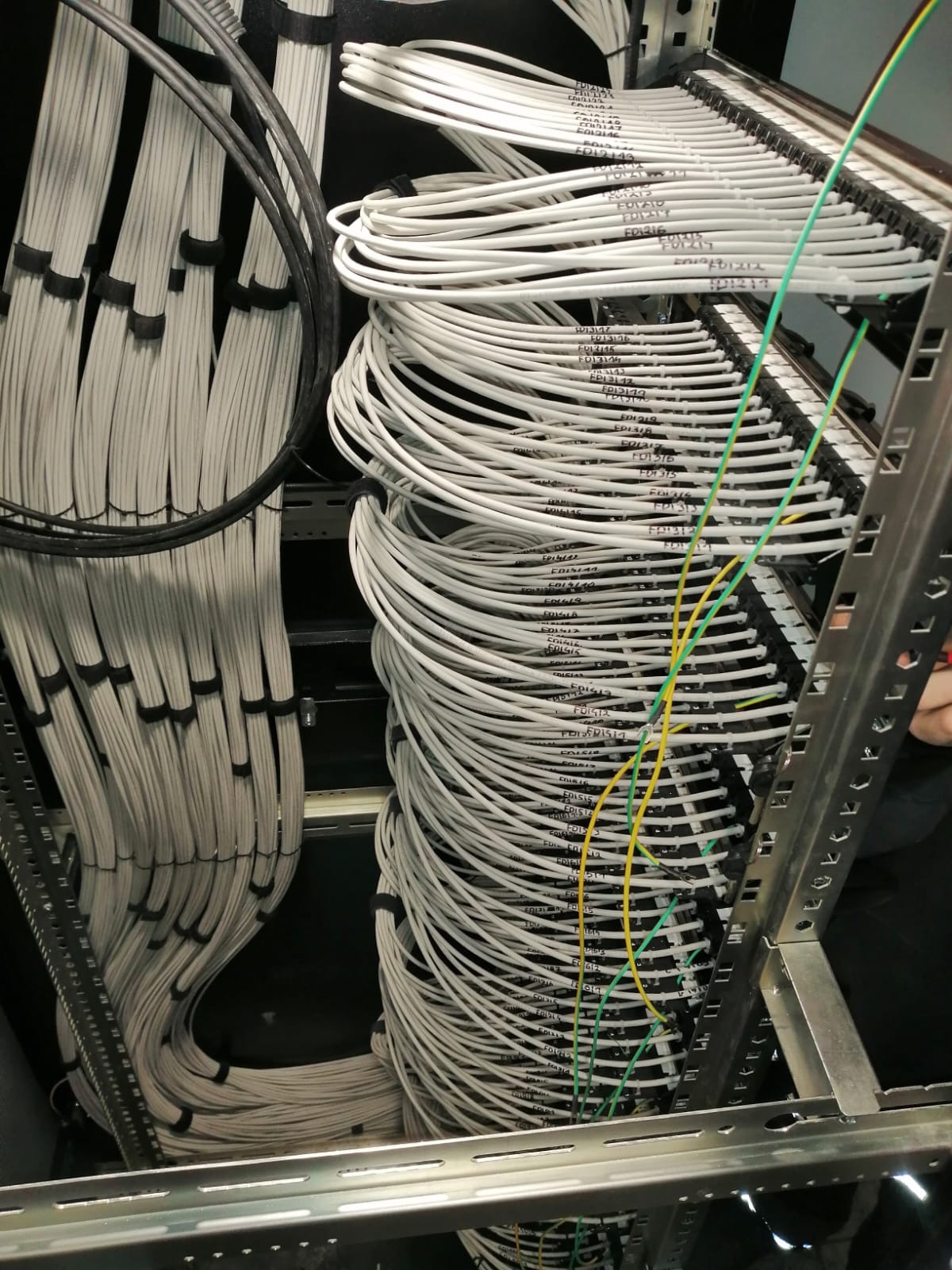 Uporządkowane kable w serwerowni, ułożone w wiązki i przymocowane do metalowej ramy. Widoczne oznaczenia na kablach i fragment dłoni w prawym górnym rogu.