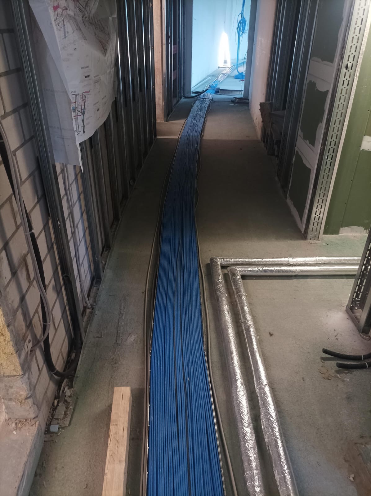 Korytarz w trakcie prac instalacyjnych: wiązka niebieskich kabli ułożona wzdłuż ściany, widoczne rury w izolacji i metalowe profile konstrukcyjne.