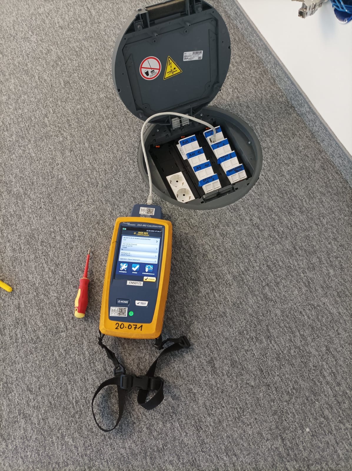 Otwarta podłogowa puszka instalacyjna z gniazdami i modułami, obok tester okablowania Fluke Networks i śrubokręt na szarym dywanie.