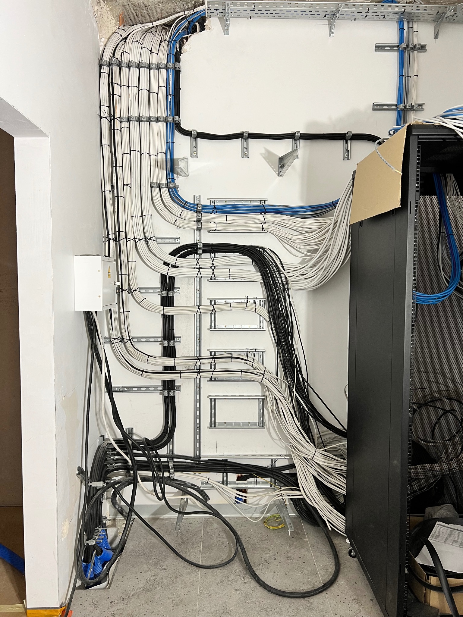 Uporządkowane kable elektryczne i telekomunikacyjne w korytach na ścianie, z szafą rack po prawej stronie. Widoczna skrzynka elektryczna i fragment surowego muru.