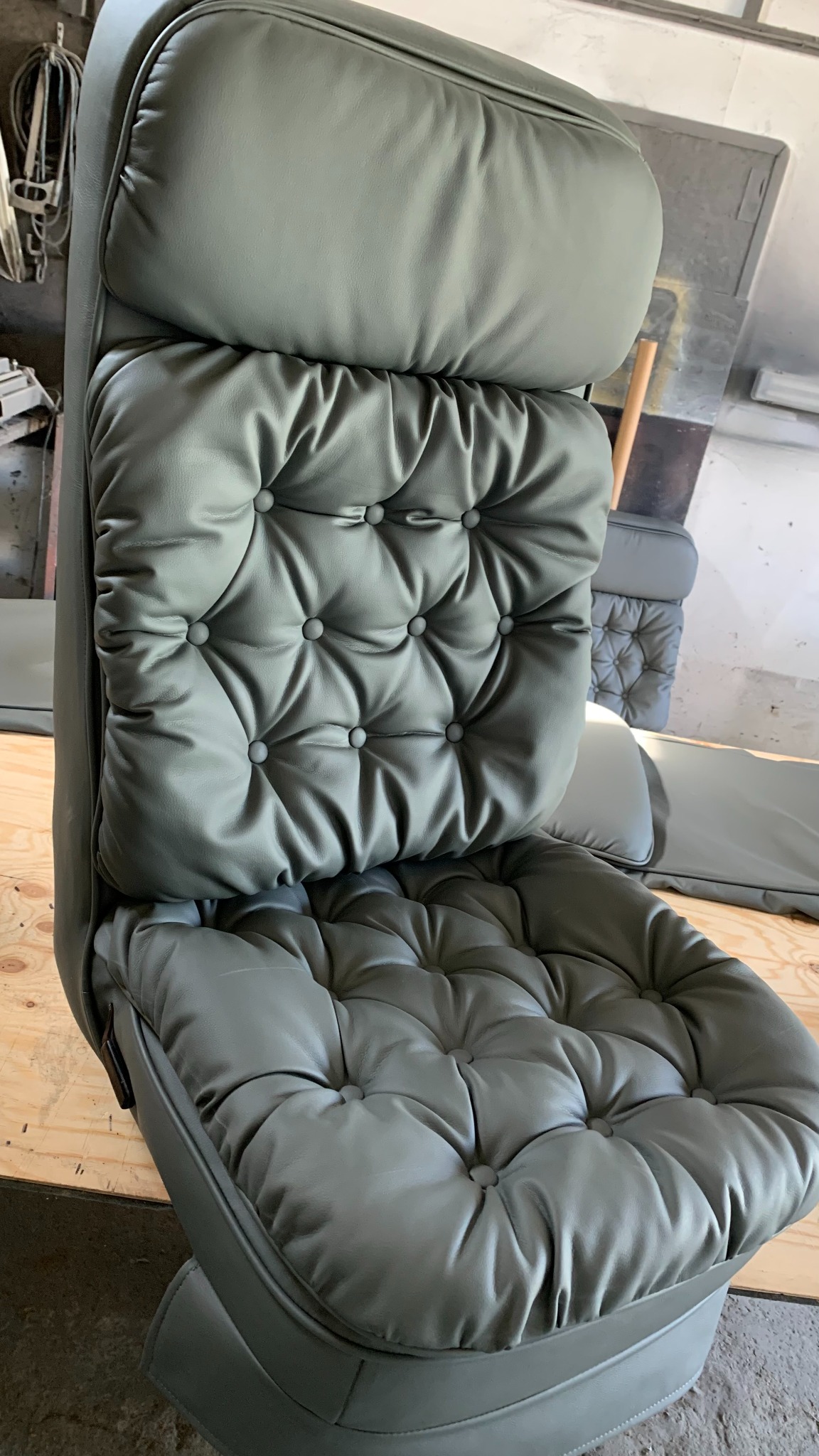 Odnowione, szare, pikowane siedzenie samochodowe z guzikami, widoczne w warsztacie. Detal tapicerki, precyzyjne wykonanie. Inne siedzenia w tle.