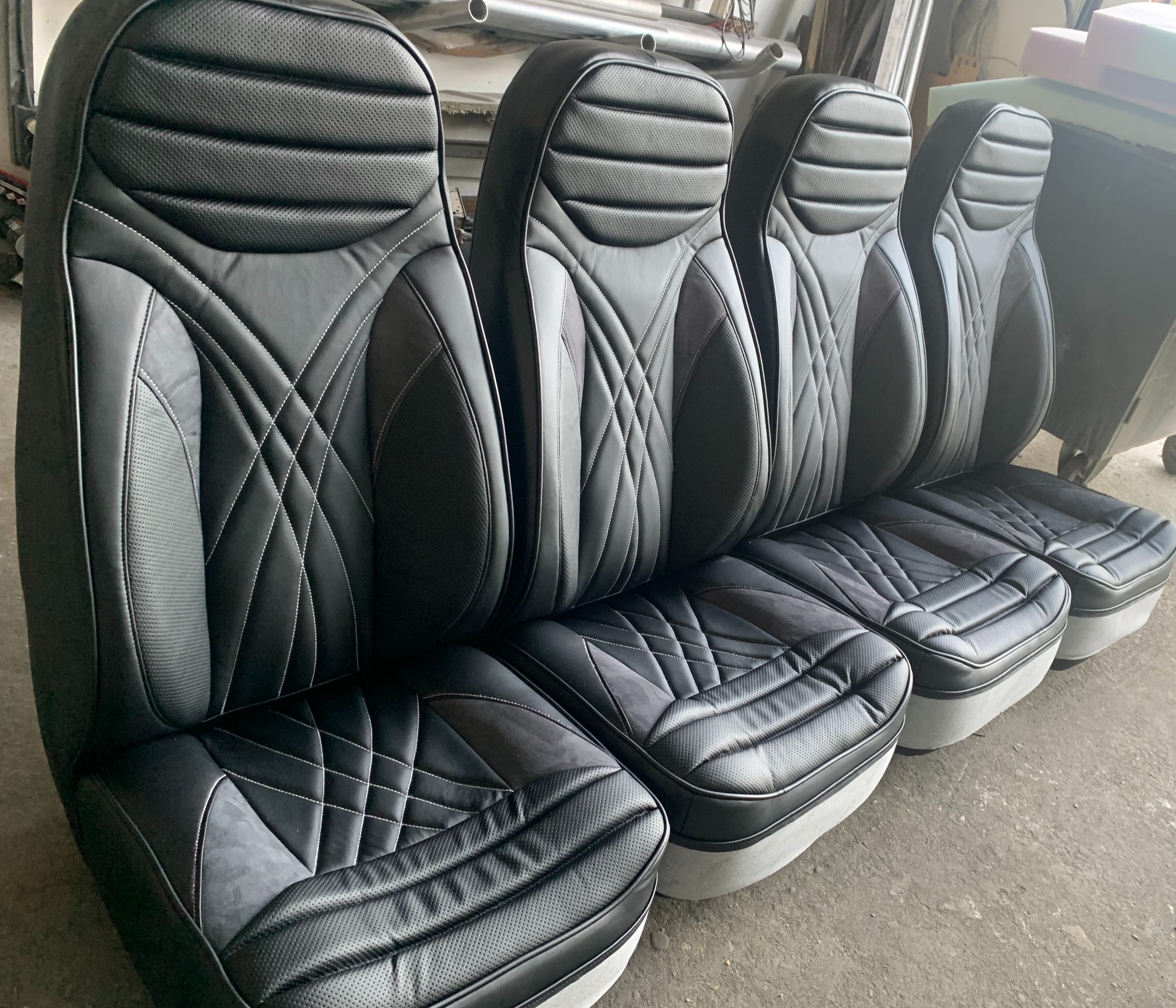 Cztery czarne, skórzane fotele samochodowe z perforowanymi wstawkami i białymi przeszyciami, ustawione obok siebie w warsztacie. Widoczne elementy metalowe w tle.