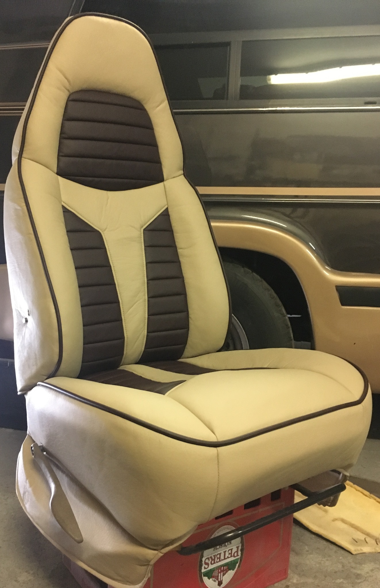 Odnowione kremowo-brązowe siedzenie samochodowe z wyraźnymi przeszyciami, umieszczone na czerwonej skrzynce, z fragmentem auta w tle. Detal tapicerki.