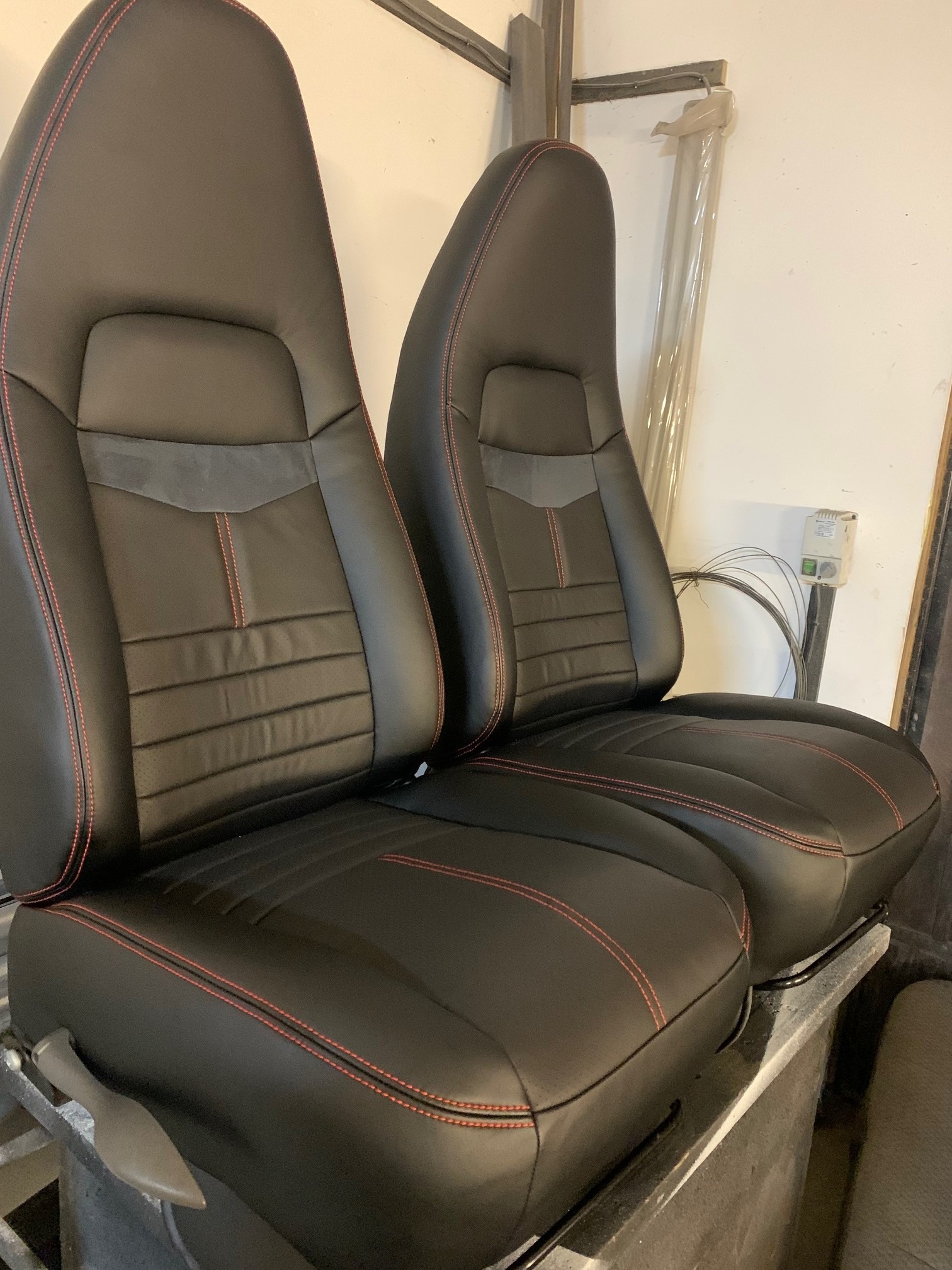 Dwa czarne, skórzane fotele samochodowe z czerwonymi przeszyciami, ustawione obok siebie. Widoczne detale wykończenia i faktura materiału.