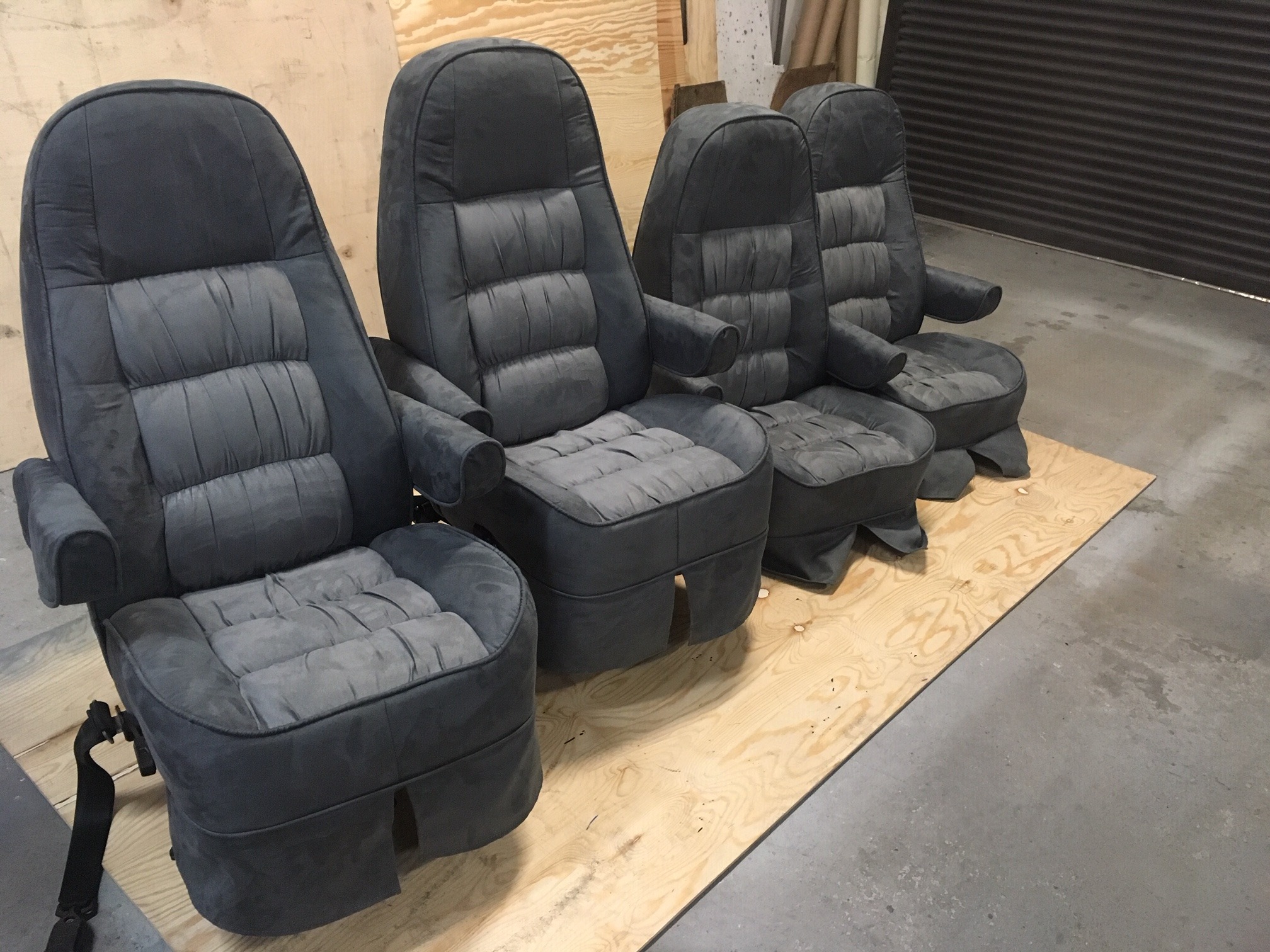 Cztery odnowione, szare fotele samochodowe z podłokietnikami, ustawione rzędem na płycie OSB w warsztacie. Widoczne detale tapicerki i pas bezpieczeństwa.