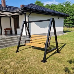 Borys Sanchuk - Metalowa huśtawka ogrodowa z drewnianym siedziskiem na tle domu z tarasem i zielonego trawnika. Czarna konstrukcja huśtawki, jasne drewno siedziska. Słoneczny dzień.