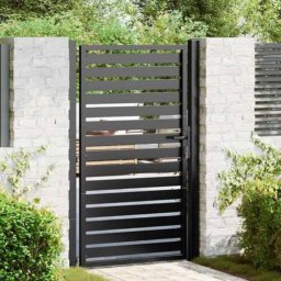 Borys Sanchuk - Nowoczesna, pozioma brama wjazdowa z czarnych profili, osadzona w białym, ceglanym murze. Minimalistyczny design, zieleń w tle.