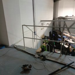 Borys Sanchuk - Biała, prosta metalowa barierka na antresoli, z której widać przestrzeń biurową z komputerami. Na betonowej podłodze leży wiertarka i kable, a z góry zwisają łańcuchy.