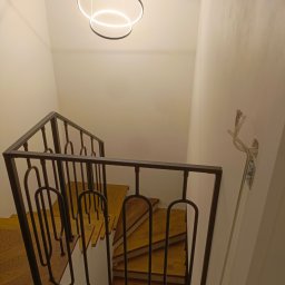 Borys Sanchuk - Widok z góry na drewniane schody z parkietem i ciemną, metalową balustradą z dekoracyjnymi, owalnymi elementami, oświetlone nowoczesnymi lampami pierścieniowymi.