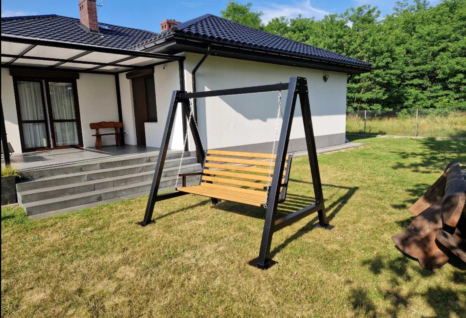 Metalowa huśtawka ogrodowa z drewnianym siedziskiem na tle domu z tarasem i zielonego trawnika. Czarna konstrukcja huśtawki, jasne drewno siedziska. Słoneczny dzień.