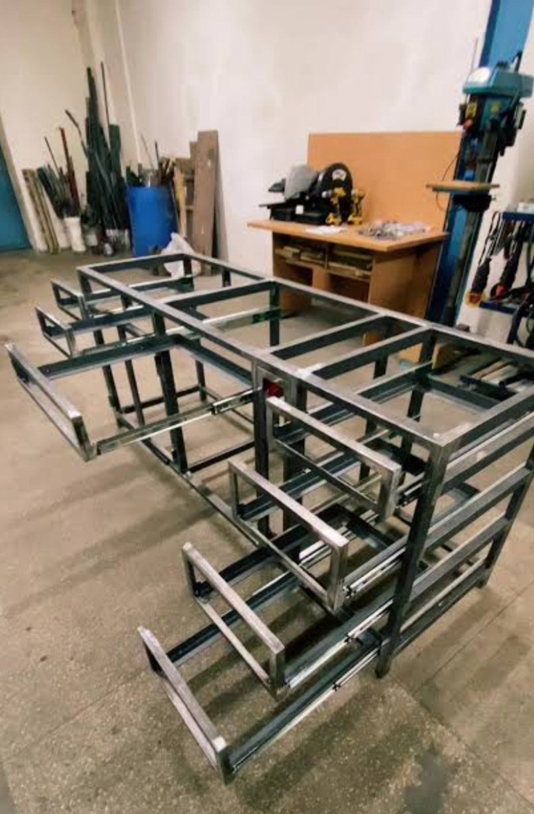 Szkielet metalowej konstrukcji z wysuwanymi elementami, prawdopodobnie ramy meblowej, w warsztacie z narzędziami w tle. Surowy, industrialny wygląd.