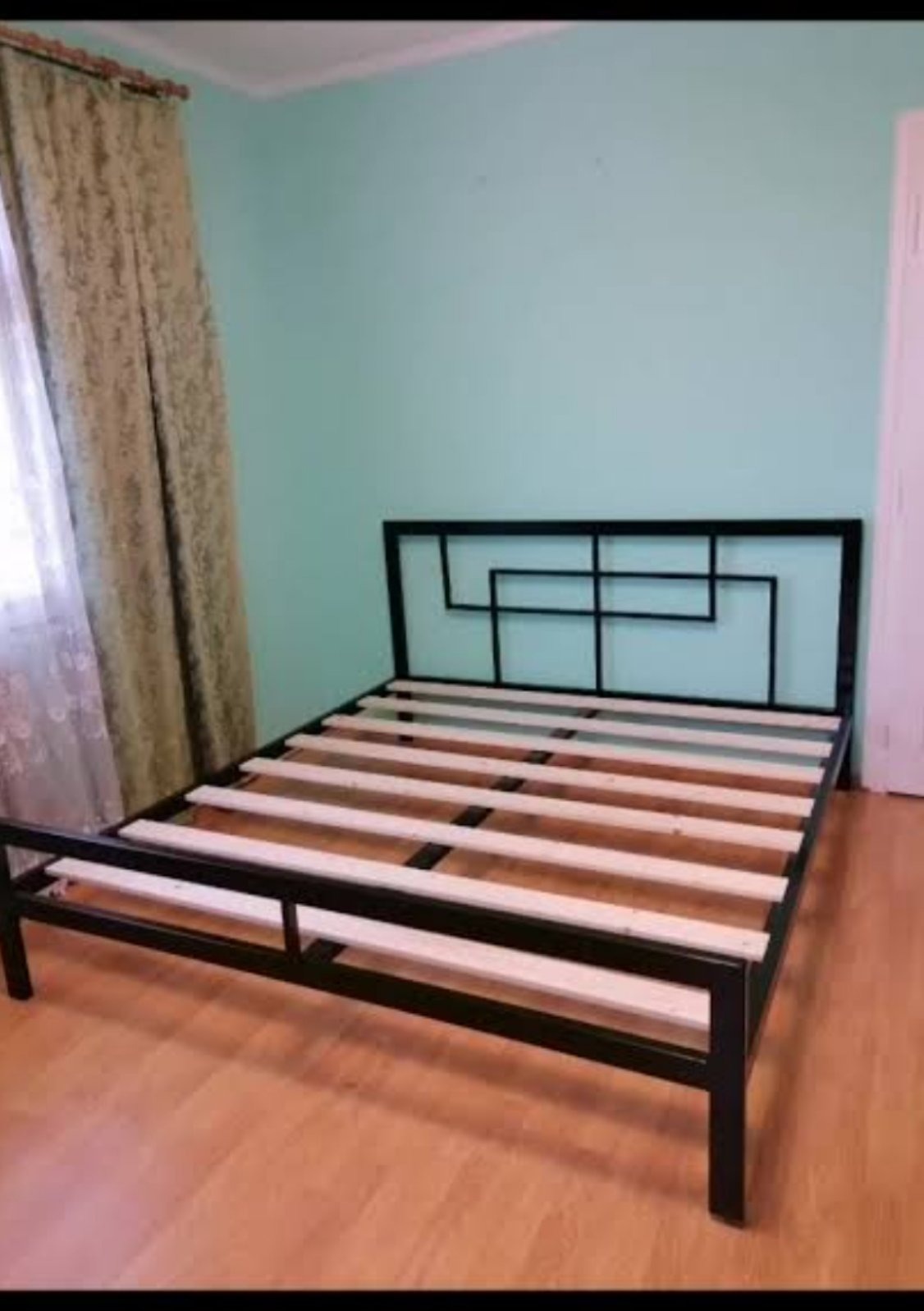 Czarna, metalowa rama łóżka z drewnianymi listwami na podłodze. Geometryczny wzór wezgłowia. Minimalistyczny design w jasnym wnętrzu.