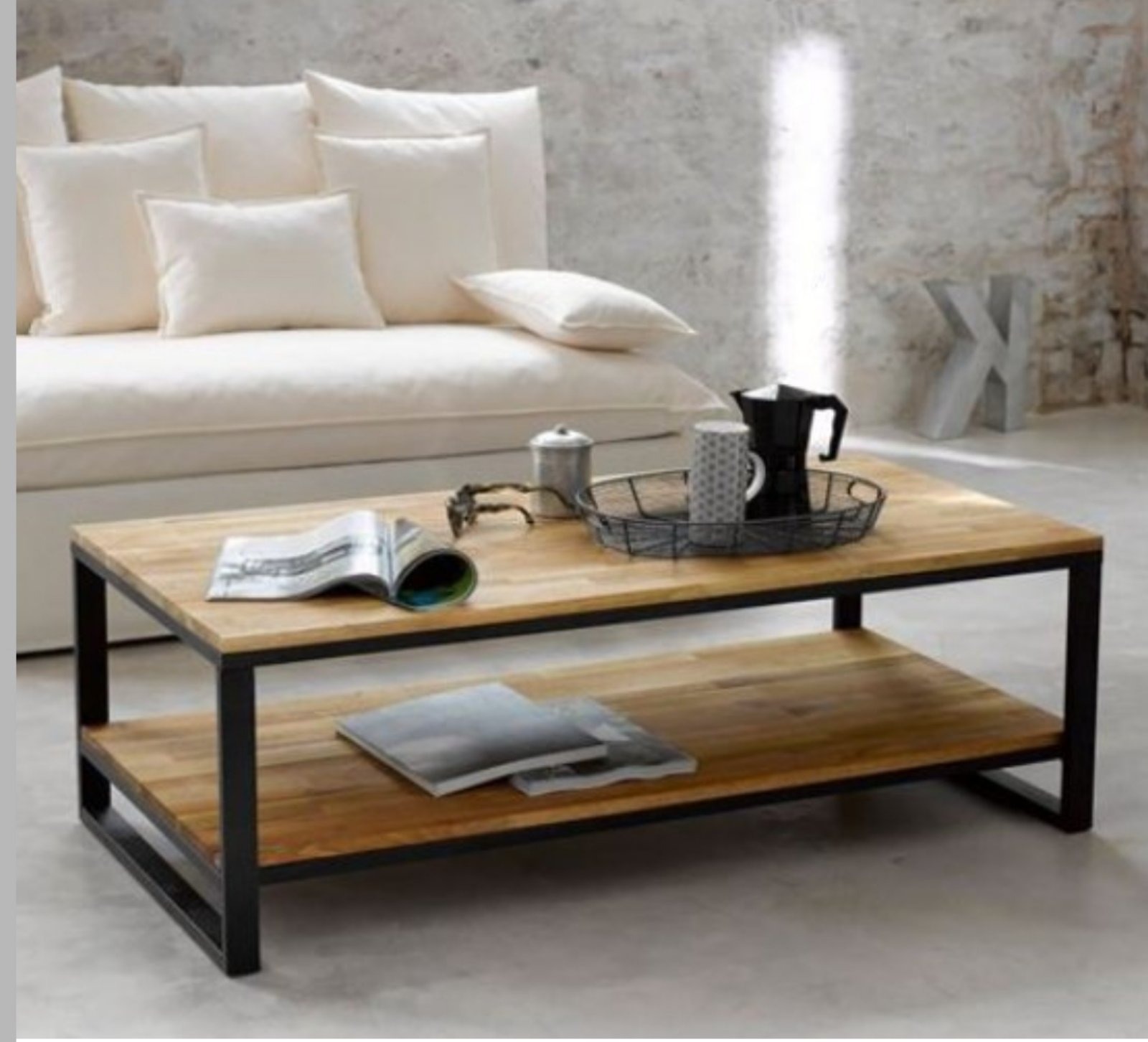 Stylowy stolik kawowy z drewnianym blatem i czarną, metalową ramą, na tle sofy z poduszkami i surowej ściany. Na blacie gazeta, okulary i akcesoria do kawy.