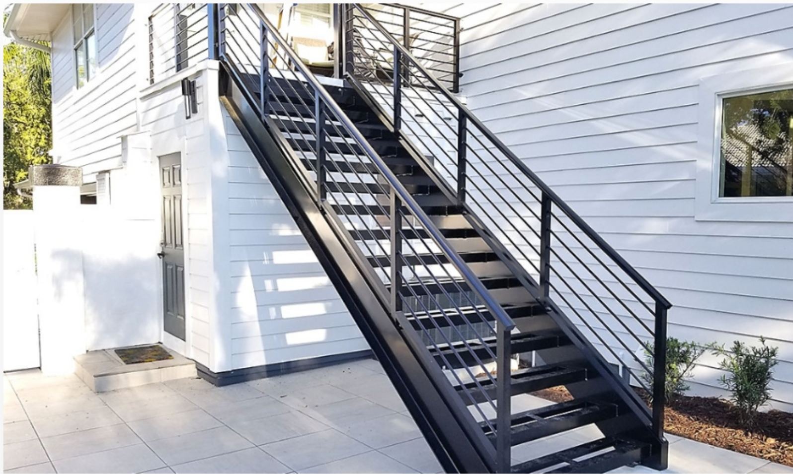 Zewnętrzne, czarne schody z metalu z poręczami, prowadzące do wejścia na piętro budynku z białą elewacją. Minimalistyczny design, jasne otoczenie.