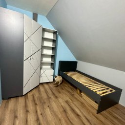 Pan od remontów - Pokój na poddaszu z drewnianą podłogą, szarą szafą z białymi frontami, regałem i łóżkiem ze stelażem. Kot przechadza się po podłodze. Ściany w odcieniach szarości i błękitu.