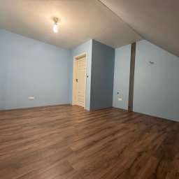 Pan od remontów - Pokój na poddaszu po remoncie: świeżo pomalowane na błękitno ściany, ułożone panele podłogowe w odcieniach brązu, białe drzwi i grzejnik. Widok na skos dachu.