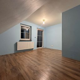 Pan od remontów - Pokój na poddaszu po odświeżeniu: drewniana podłoga, błękitne ściany, okno z grzejnikiem. Widoczne skosy i surowe wykończenie sufitu.