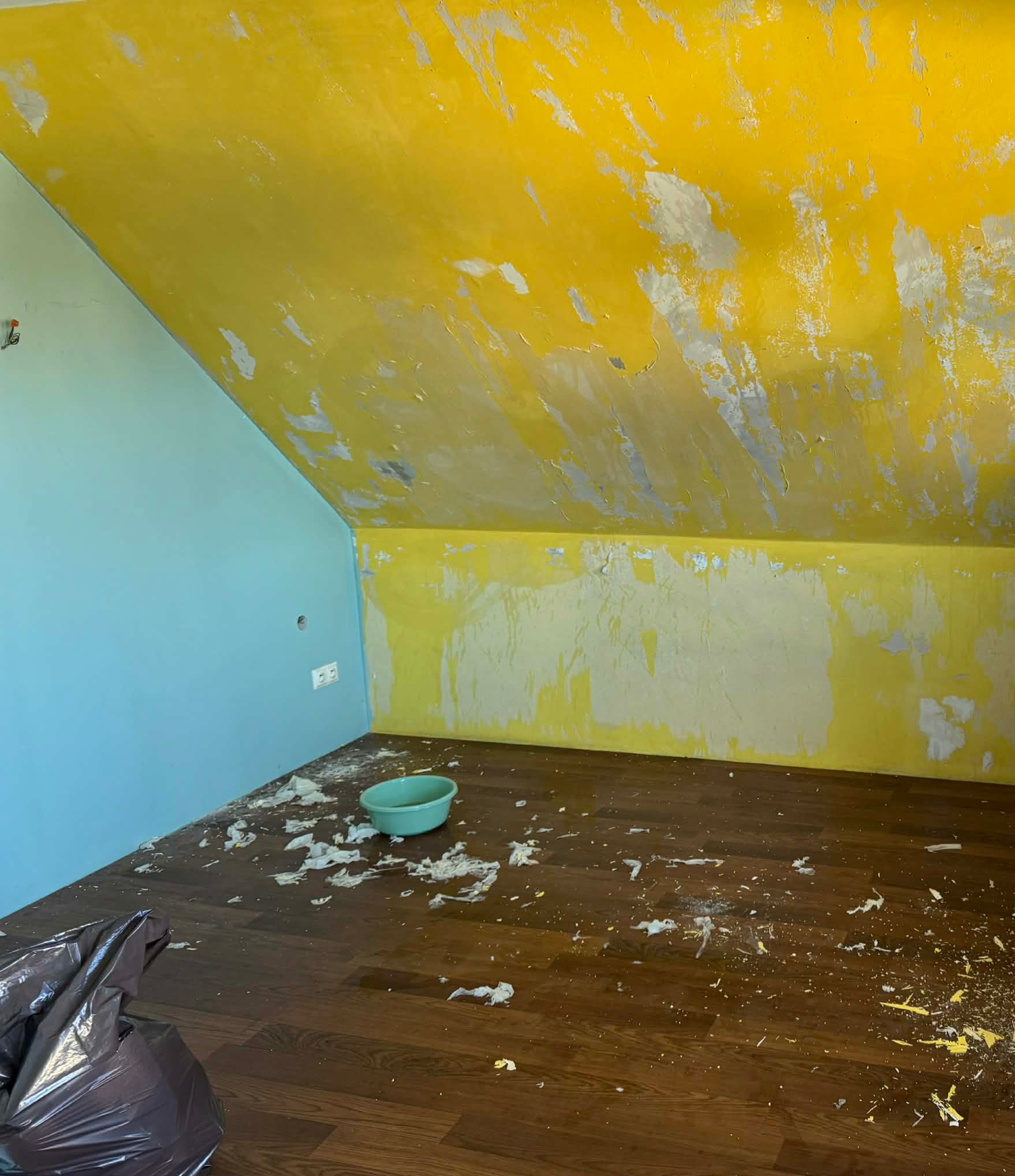 Pokój na poddaszu w trakcie przygotowania do malowania: ściany częściowo ogołocone z farby, widoczne resztki farby na podłodze, worek na śmieci i miska.