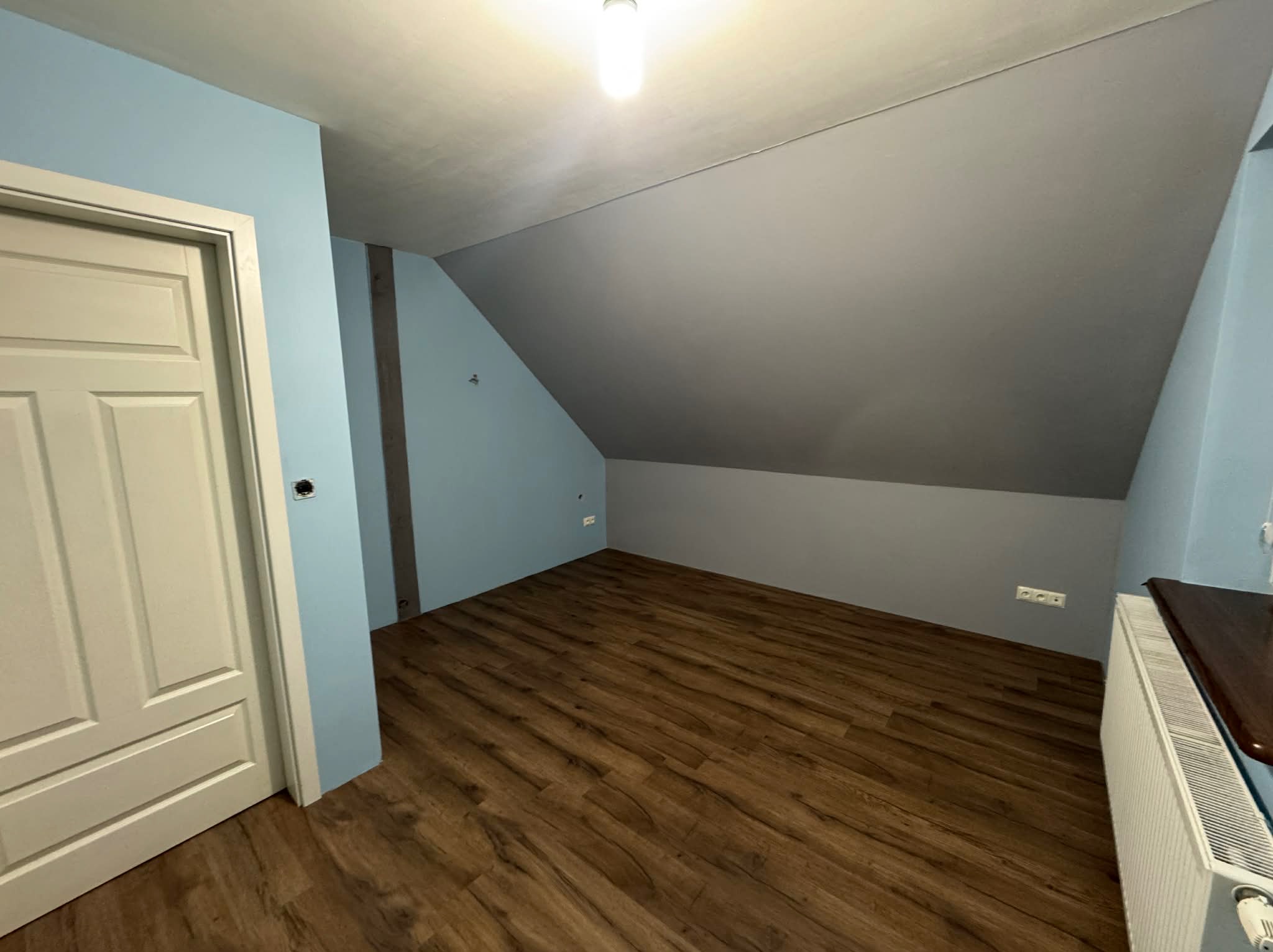 Pokój na poddaszu po remoncie: świeżo ułożone panele podłogowe, pomalowane ściany na niebiesko i szaro, białe drzwi i grzejnik. Widok na skos dachu i oświetlenie.