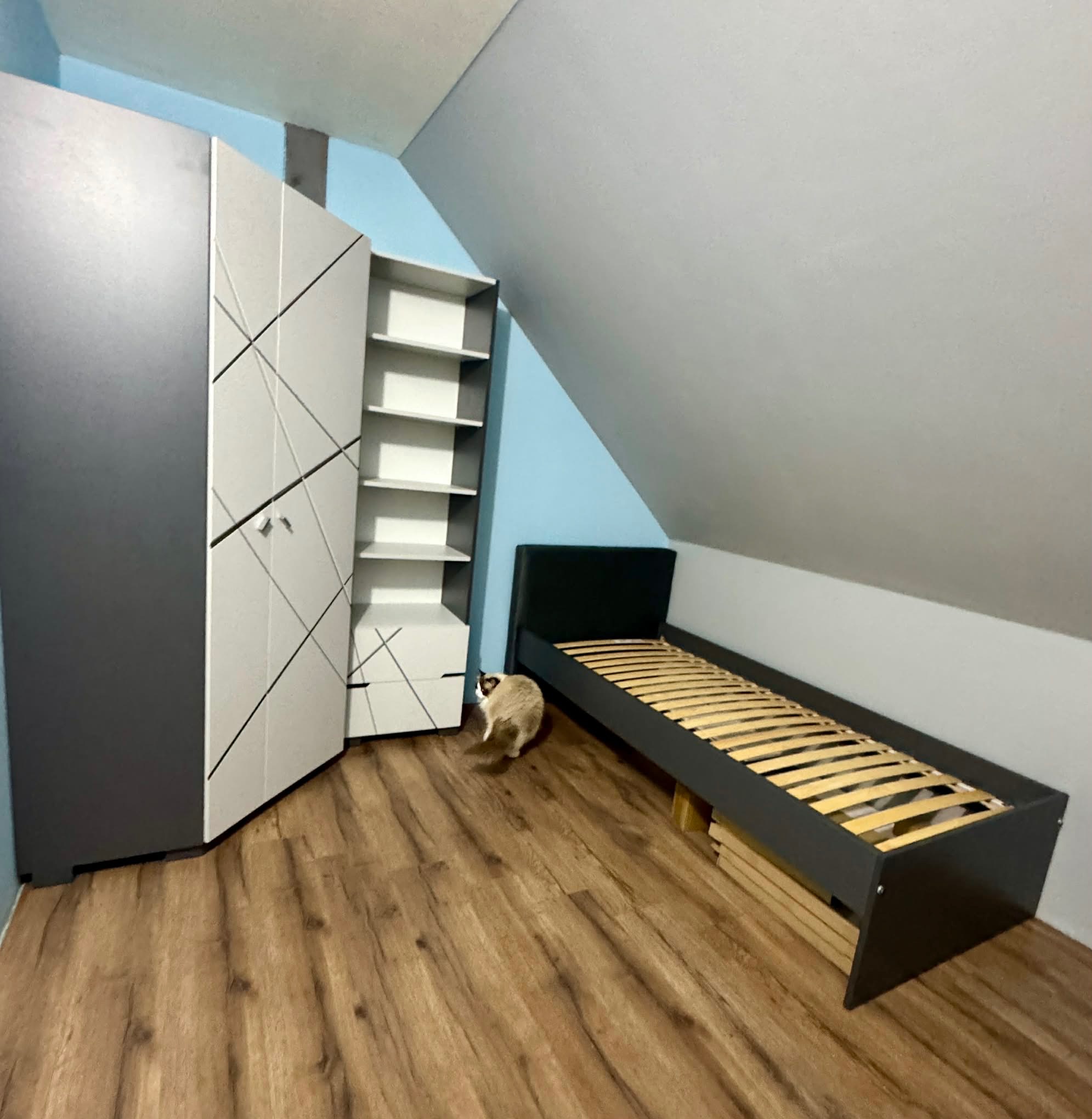 Pokój na poddaszu z drewnianą podłogą, szarą szafą z białymi frontami, regałem i łóżkiem ze stelażem. Kot przechadza się po podłodze. Ściany w odcieniach szarości i błękitu.