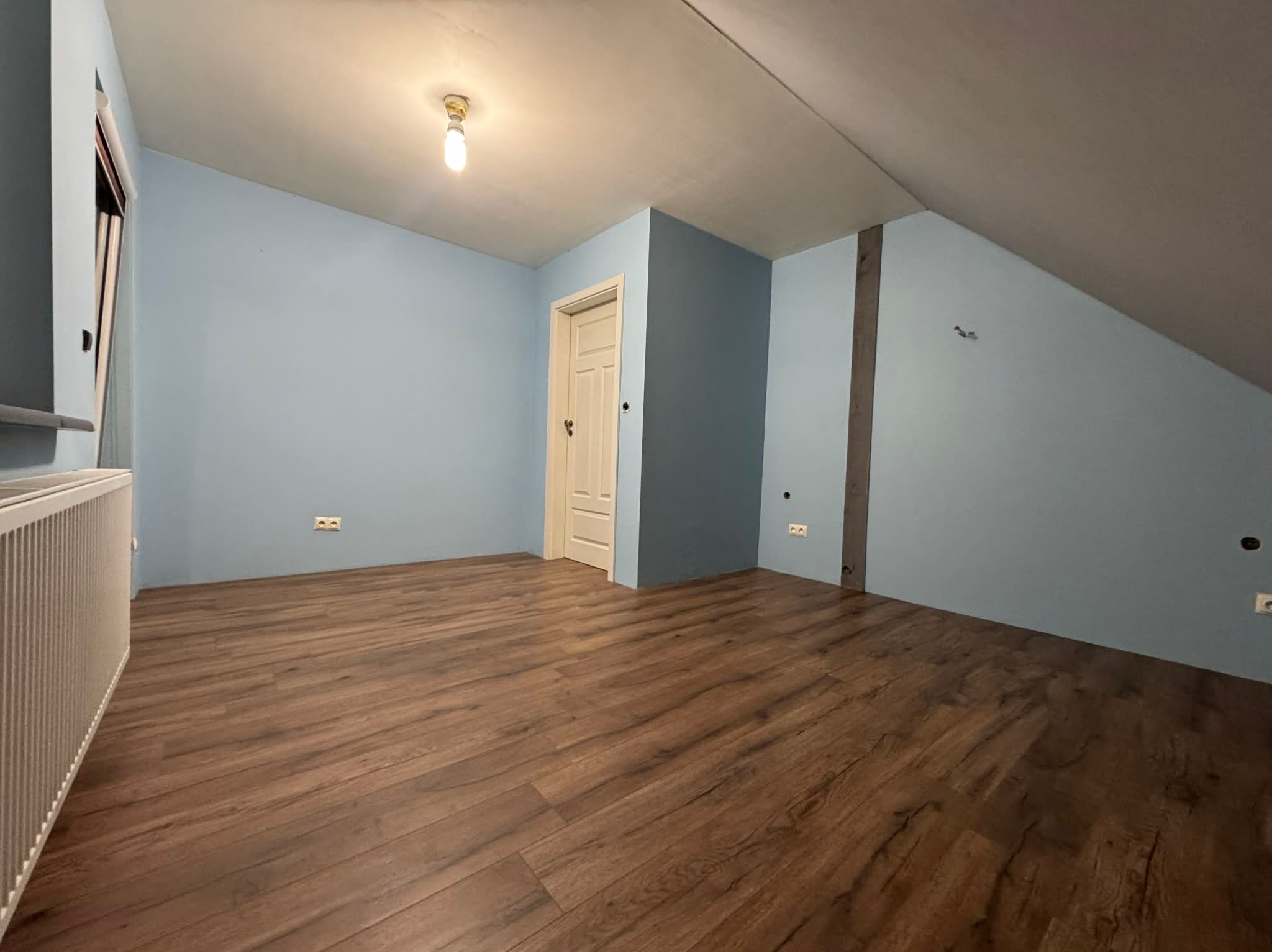 Pokój na poddaszu po remoncie: świeżo pomalowane na błękitno ściany, ułożone panele podłogowe w odcieniach brązu, białe drzwi i grzejnik. Widok na skos dachu.