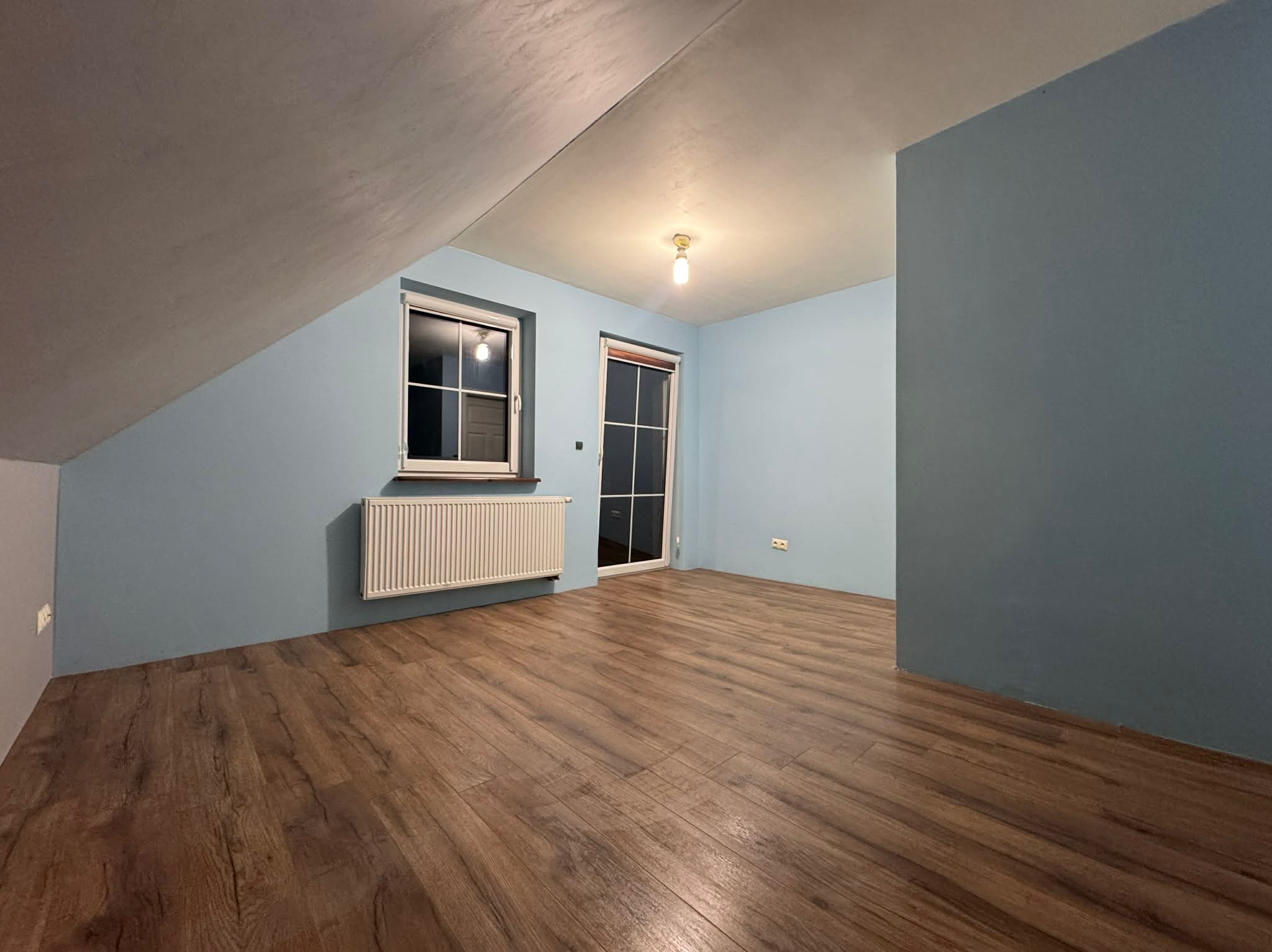 Pokój na poddaszu po odświeżeniu: drewniana podłoga, błękitne ściany, okno z grzejnikiem. Widoczne skosy i surowe wykończenie sufitu.