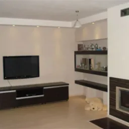 Jasne wnętrze salonu z telewizorem na ścianie, zabudową kominkową z ciemnobrązowymi akcentami, półkami i pluszowym misiem leżącym na podłodze.