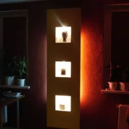 Podświetlana dekoracja ścienna z trzema kwadratowymi wnękami, zawierającymi różne obiekty dekoracyjne, na tle ciemnoczerwonej ściany z widoczną fakturą.