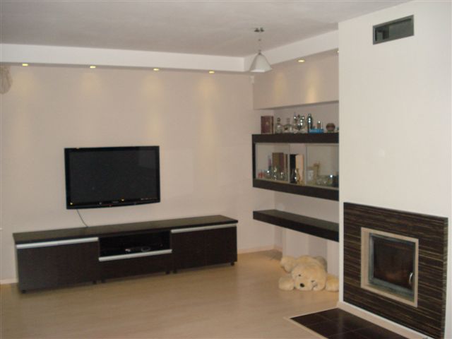 Jasne wnętrze salonu z telewizorem na ścianie, zabudową kominkową z ciemnobrązowymi akcentami, półkami i pluszowym misiem leżącym na podłodze.