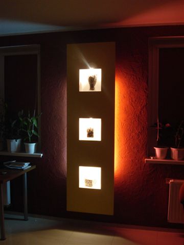 Podświetlana dekoracja ścienna z trzema kwadratowymi wnękami, zawierającymi różne obiekty dekoracyjne, na tle ciemnoczerwonej ściany z widoczną fakturą.