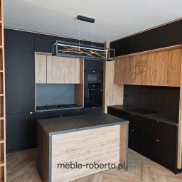 Meble roberto - Nowoczesna kuchnia z wyspą: czarne fronty, drewniane akcenty, marmurowy blat. Oświetlenie LED w geometrycznej lampie. Widoczny regał z jasnego drewna i zabudowa piekarnika.